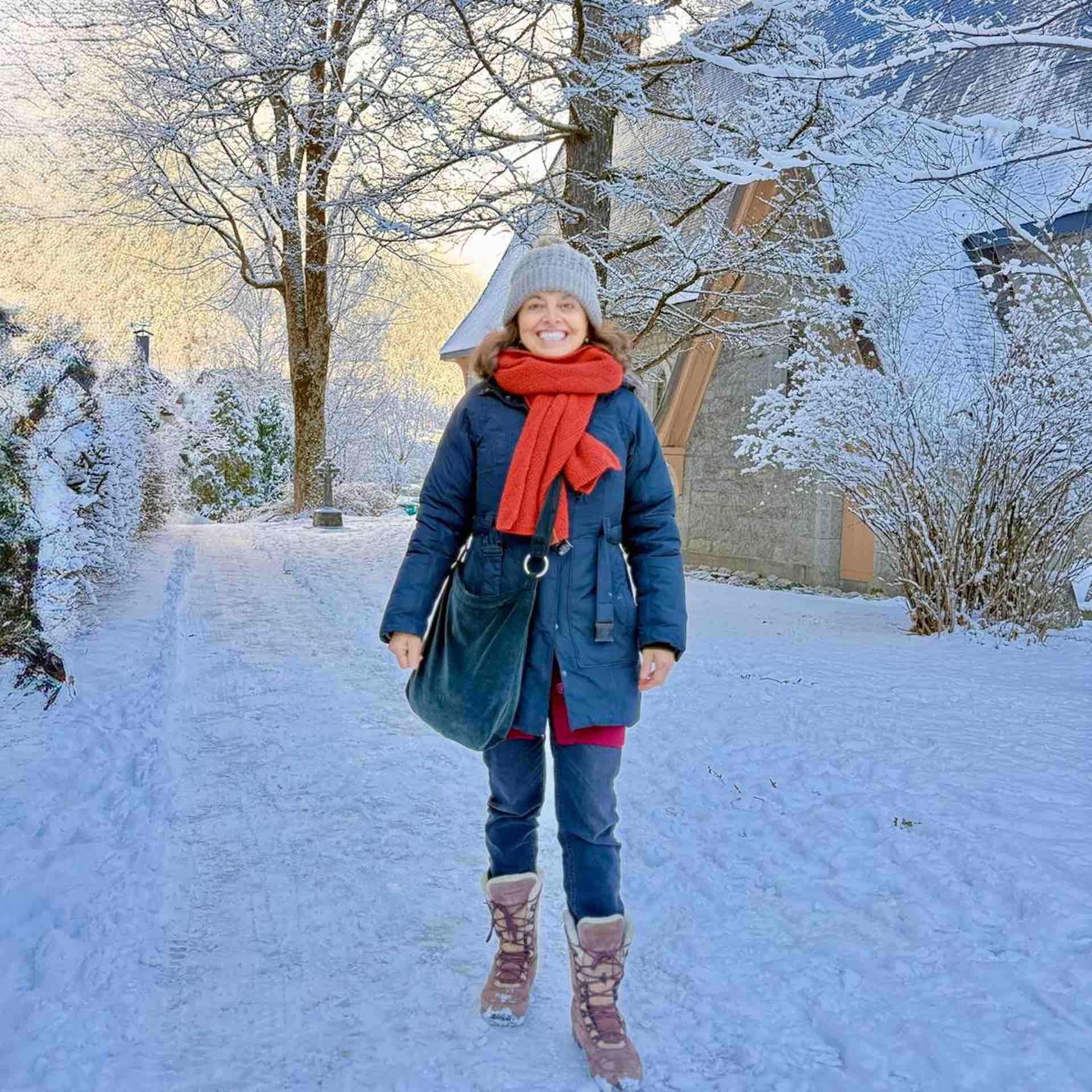 Chicca in a navy winter coat, red scarf, and grey beanie smiling while standing on a snow-covered path lined with frosted trees and a small wooden cabin in Chamonix.