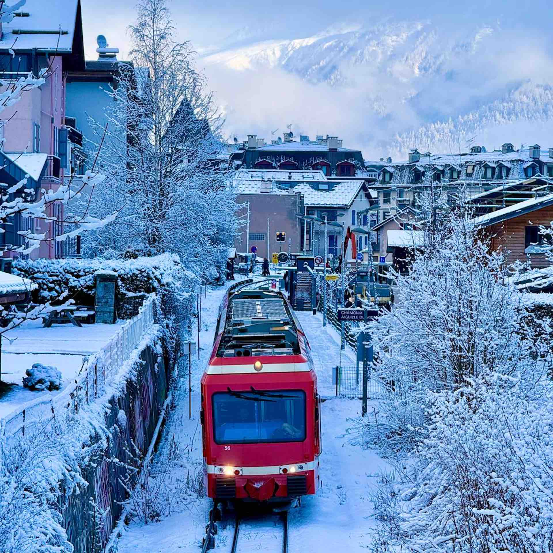 A head-on view of the iconic red Montenvers cog train traveling along snow-dusted tracks in Chamonix through a charming, wintery alpine village with mountains in the background.