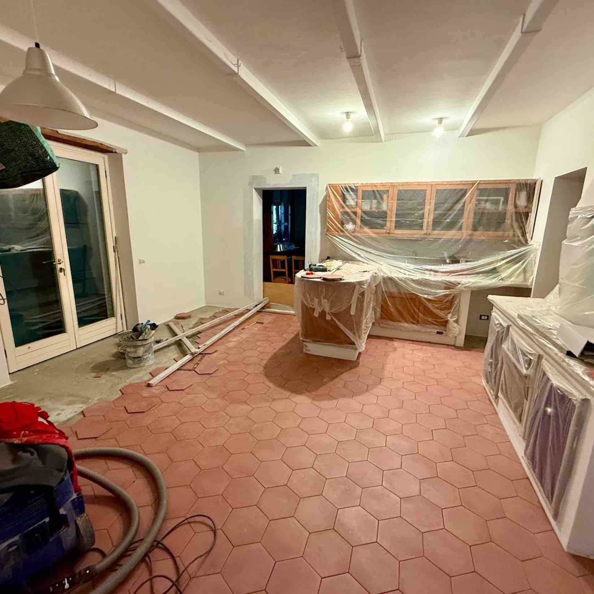 Kitchen under renovation in a Tuscan farmhouse featuring unsealed terracotta hexagon floor tiles, white beamed ceilings, and protected cabinetry.