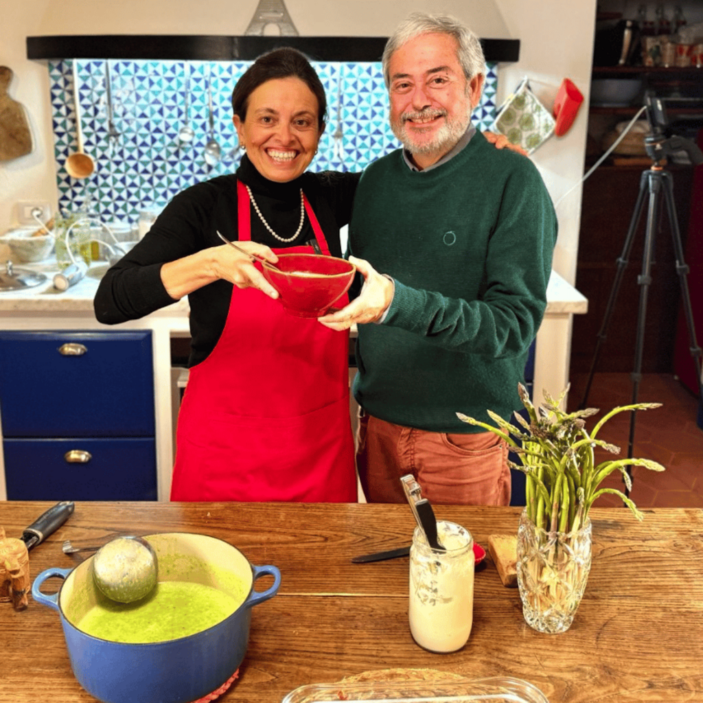 Chicca and Giuliano Hazan smile in the Tuscan farmhouse kitchen while holding a bowl of asparagus soup.