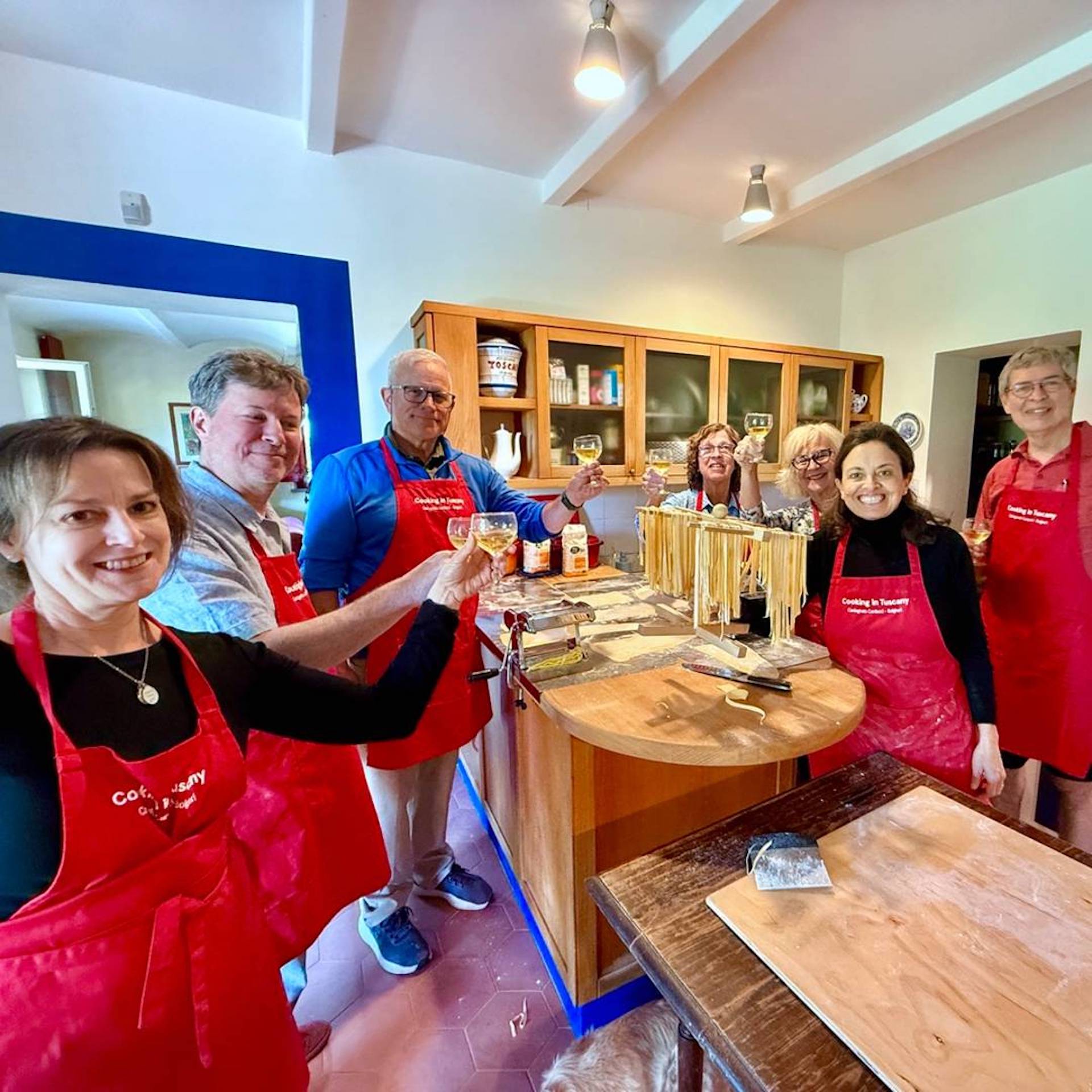 Guests in red Cooking in Tuscany aprons toast in Chicca’s Tuscan farmhouse kitchen beside freshly made pasta drying on a rack.