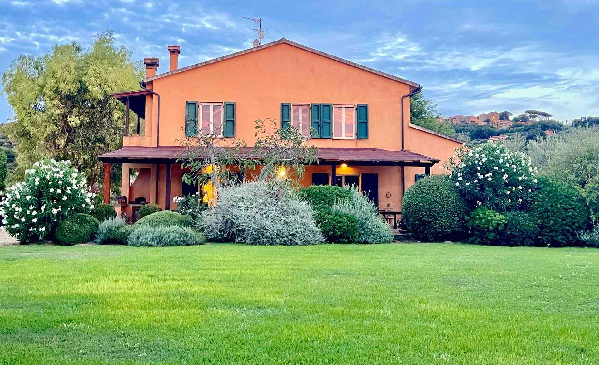 Orange Tuscan farmhouse with green shutters, surrounded by a garden and blue sky