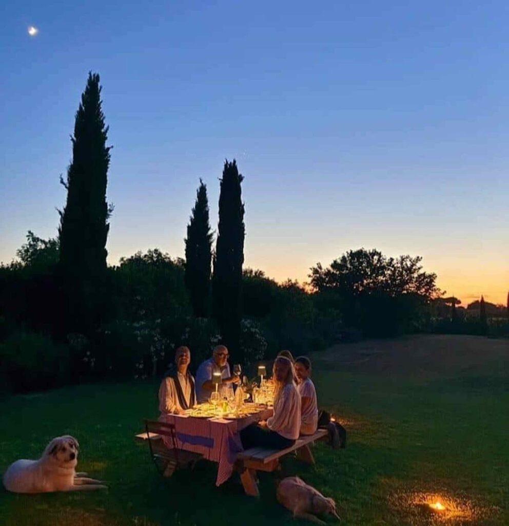 Family and dogs gathered around an outdoor dinner table at dusk beneath tall Tuscan cypress trees
