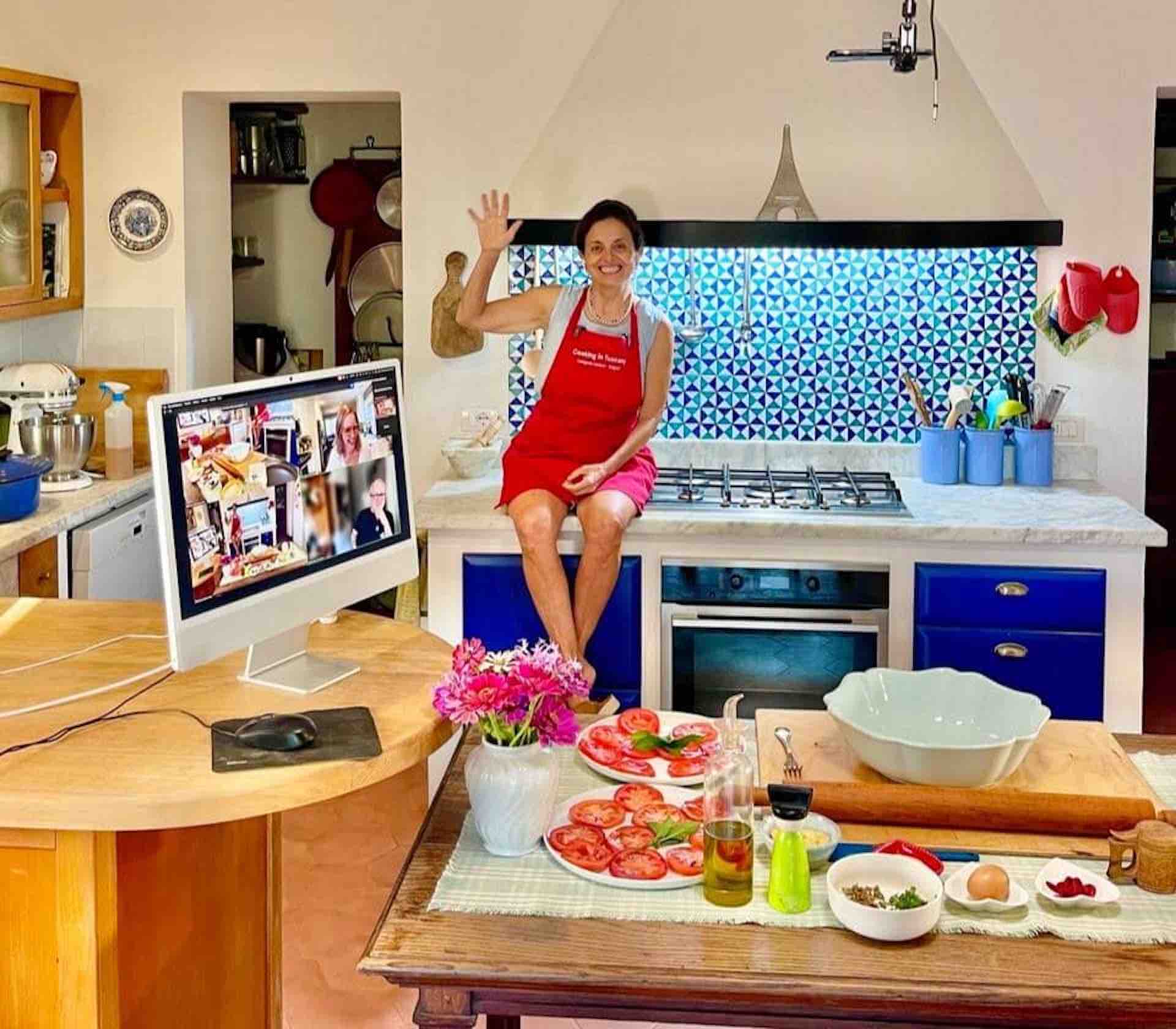 Chicca waving from her Tuscan kitchen counter beside a laptop showing class participants, with fresh tomatoes on the table
