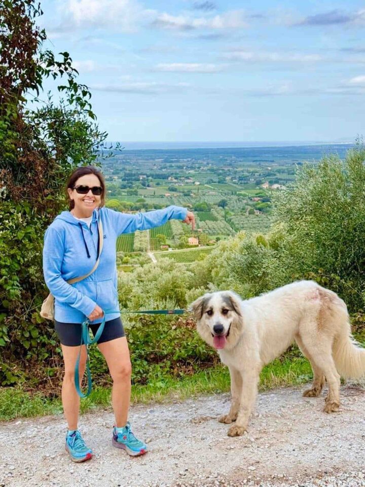 Chicca in a blue jacket and sunglasses standing on a hillside path with a large white dog, pointing toward the green Tuscan valley below