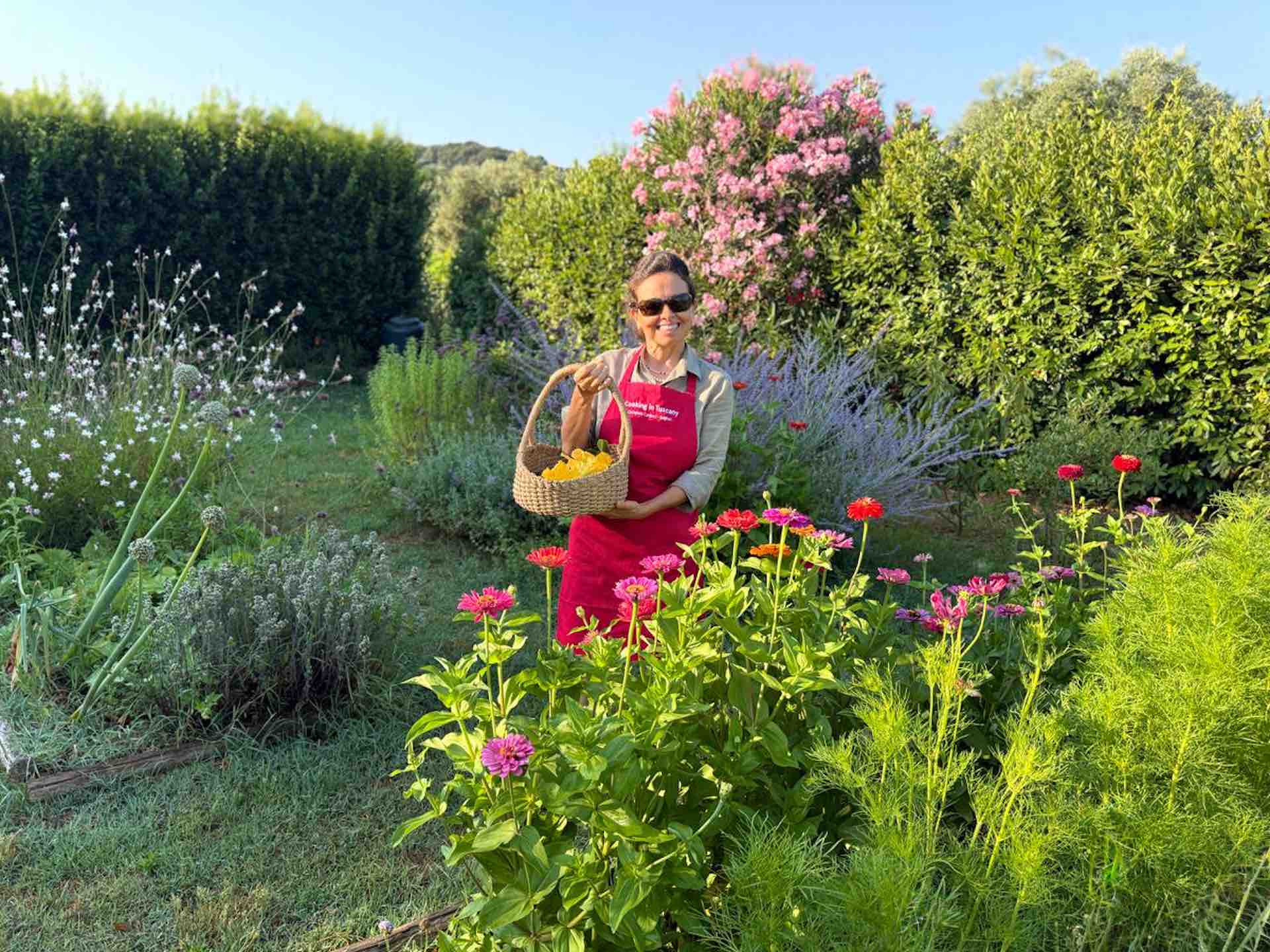 Chicca standing in a lush Tuscan garden full of flowers and herbs, holding a basket of fresh vegetables