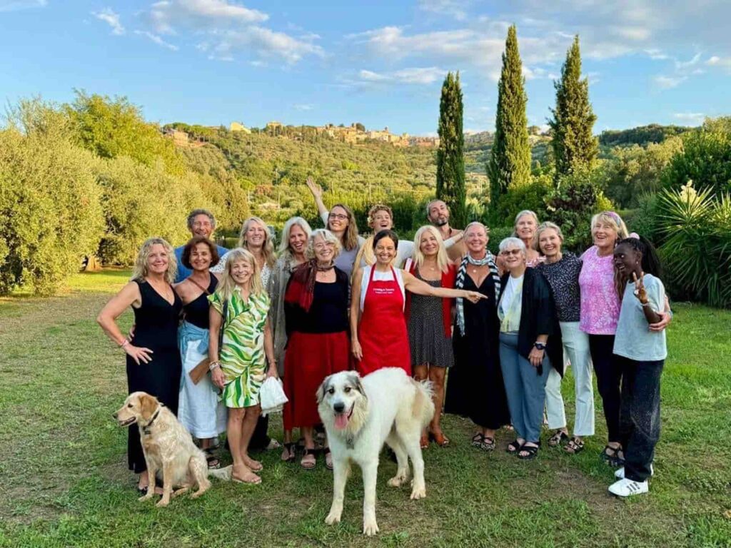 Group of smiling cooking class guests standing together in a Tuscan garden at sunset, with Chicca in a red apron and two dogs in front