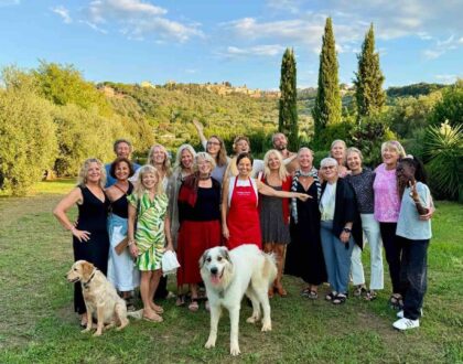 Group of smiling cooking class guests standing together in a Tuscan garden at sunset, with Chicca in a red apron and two dogs in front
