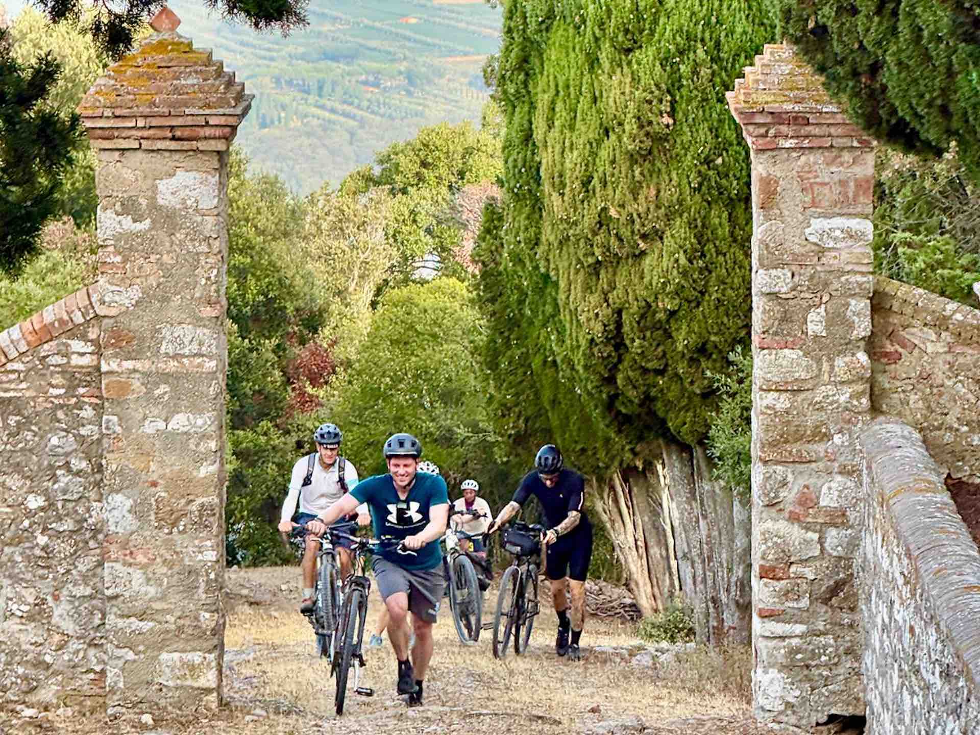 Group of cyclists riding bicycles through an old stone gate on the private road to Castiglioncello di Bolgheri