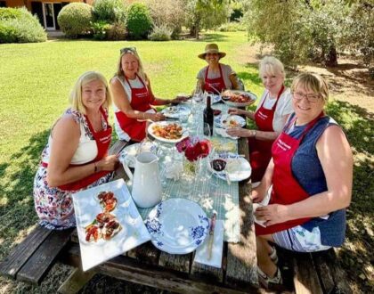 Five women in red Cooking in Tuscany aprons seated at an outdoor table with plates of food, celebrating Season 21