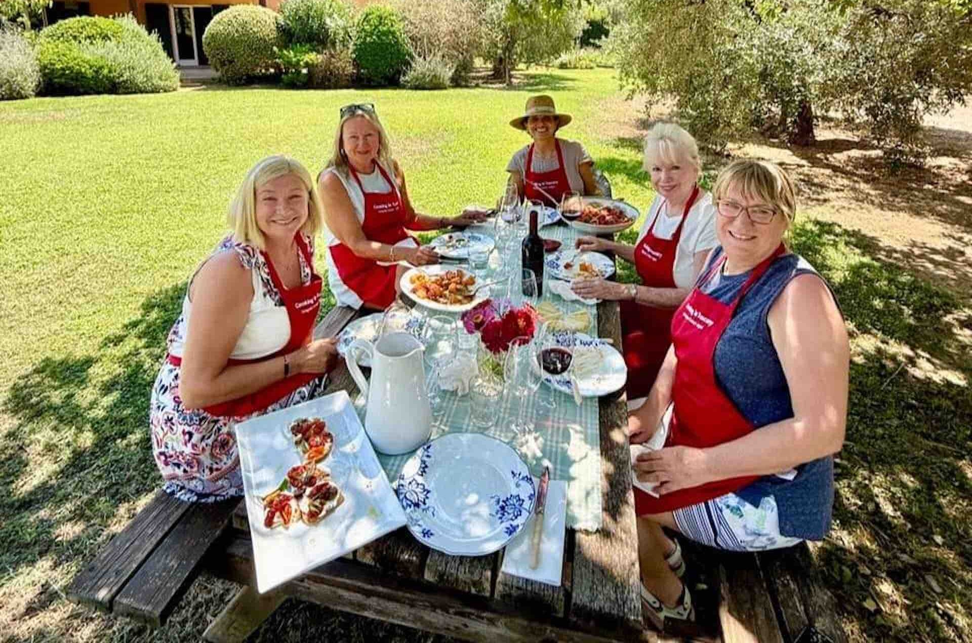 Five women in red Cooking in Tuscany aprons seated at an outdoor table with plates of food, celebrating Season 21
